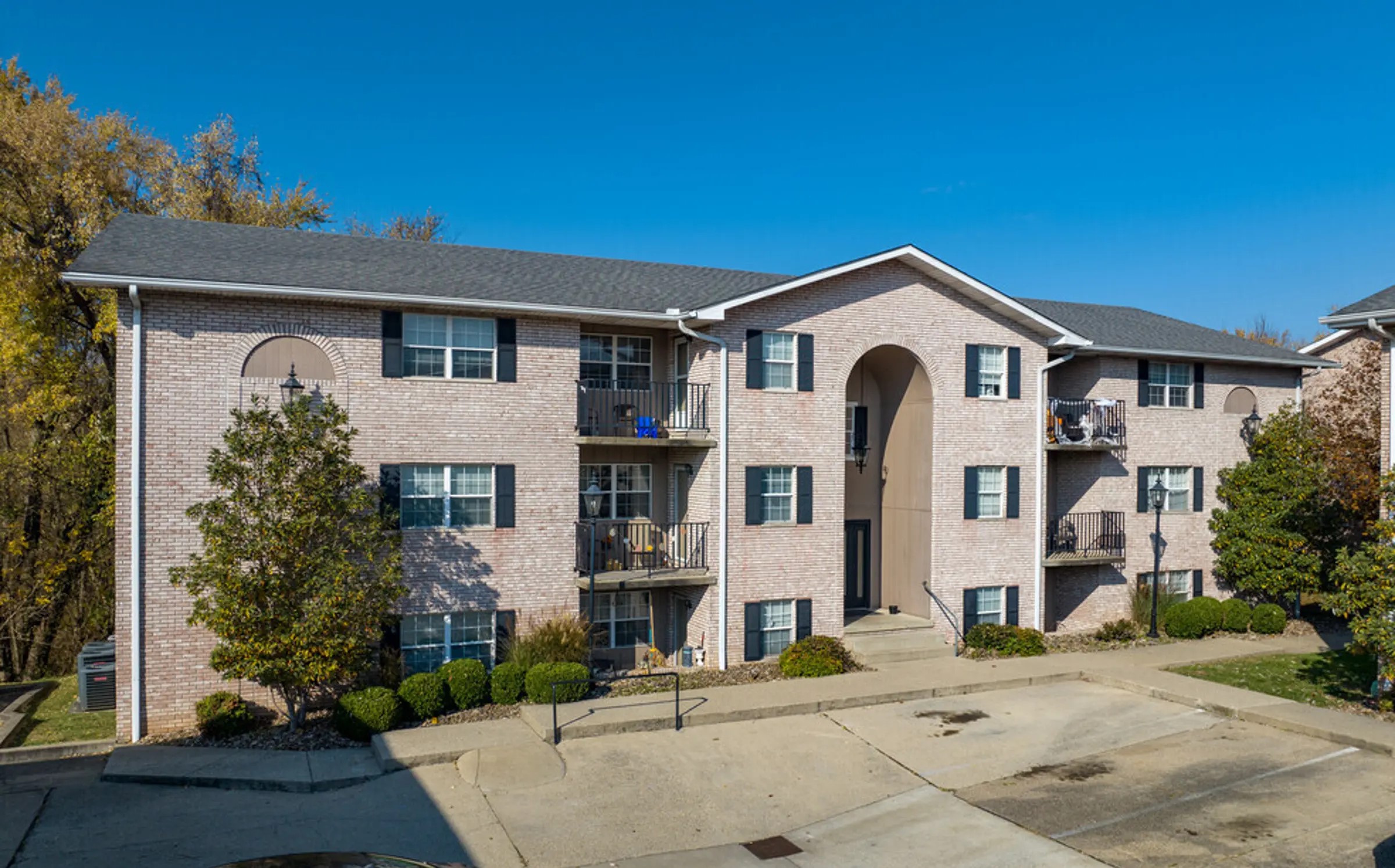 A well-maintained, three-story brick apartment building with a welcoming arched entryway and classic black shutters. The exterior is complemented by neatly trimmed landscaping, offering a sense of comfort and tranquility. The building’s balconies provide cozy spaces for relaxation, making it ideal for anyone looking for a serene, inviting place to call home. The clean, spacious parking area adds convenience, making this a highly attractive living space.